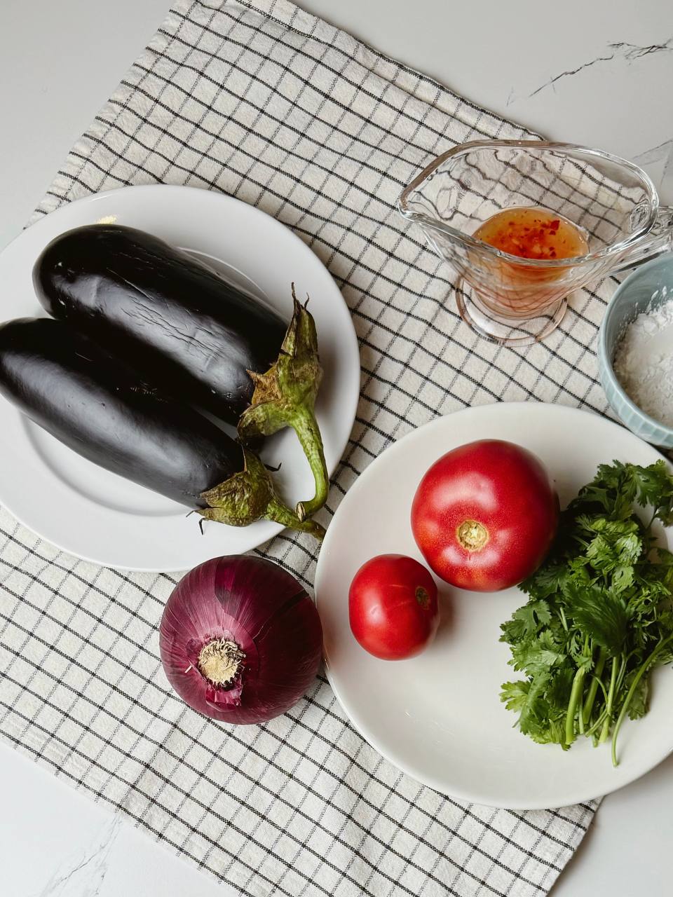 A top-down view of fresh ingredients for crispy eggplant salad, including eggplants, tomatoes, red onion, cilantro, and sweet chili sauce.