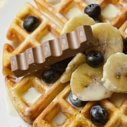 Belgian waffles topped with banana slices, condensed milk, Kinder chocolate pieces, and fresh berries.