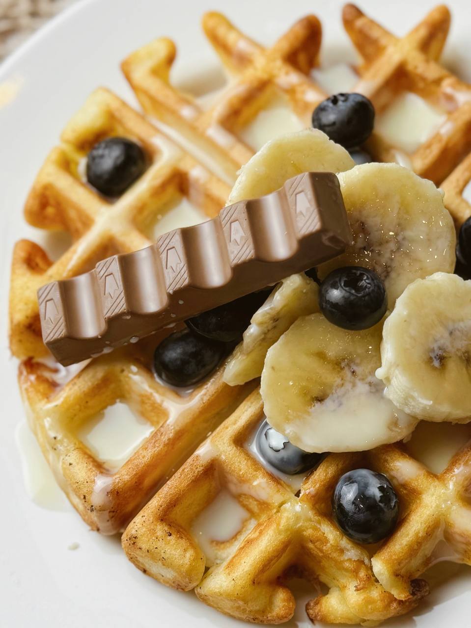 Belgian waffles topped with banana slices, condensed milk, Kinder chocolate pieces, and fresh berries.