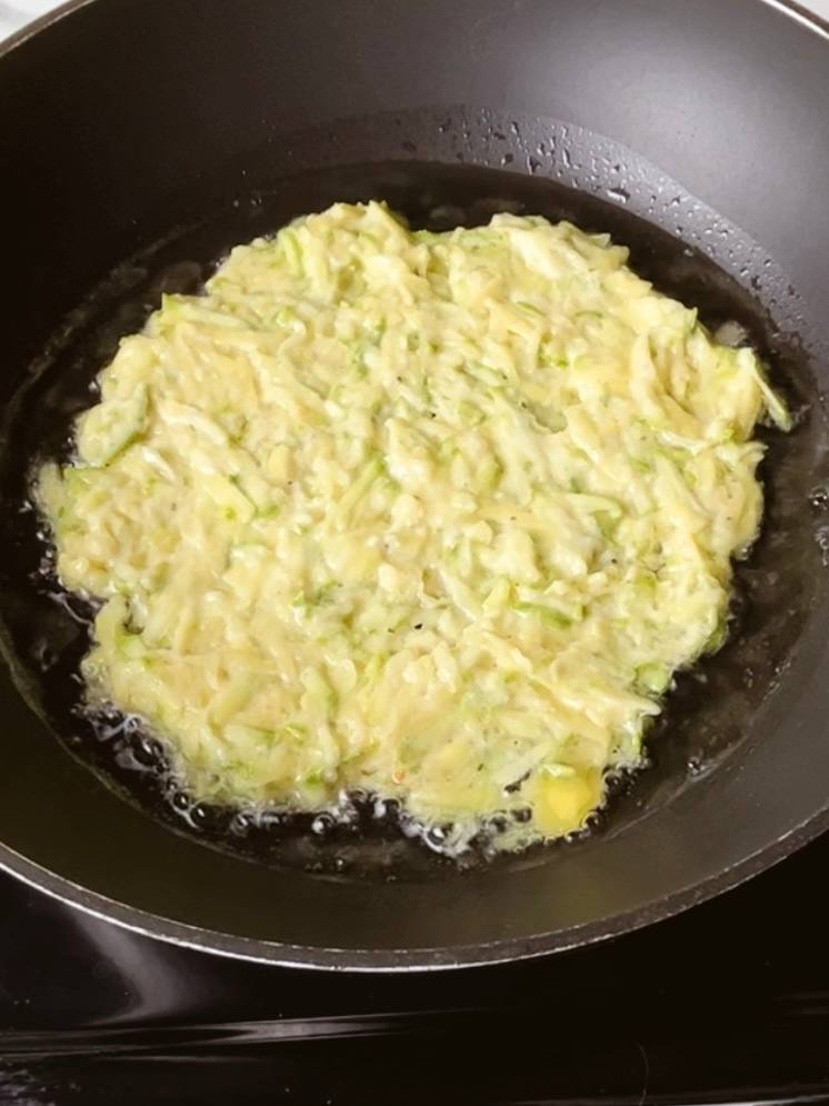 Potato zucchini pancakes spread in a frying pan, cooking until golden.