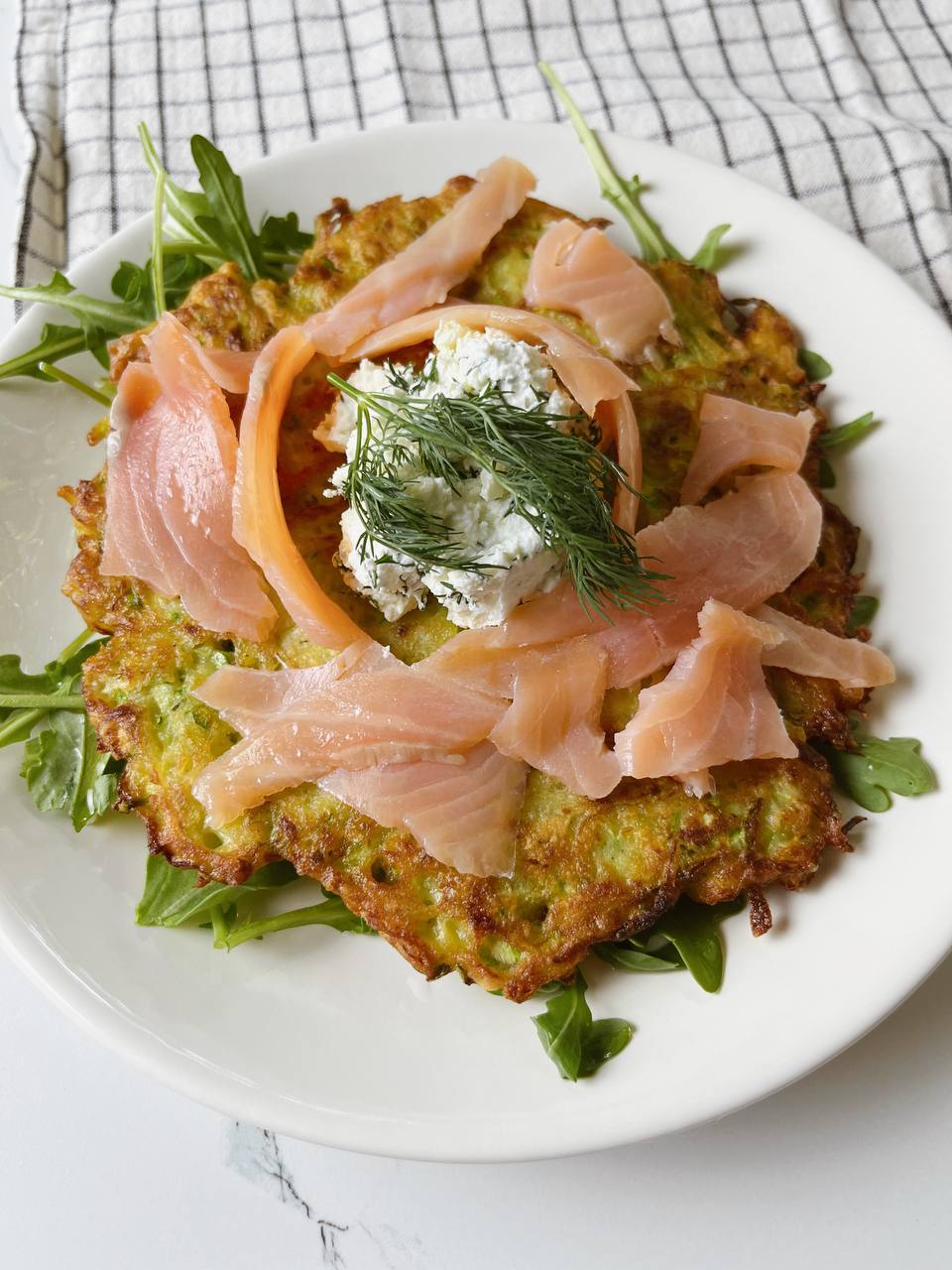 A plated potato zucchini pancake with smoked salmon, cream cheese, and arugula.
