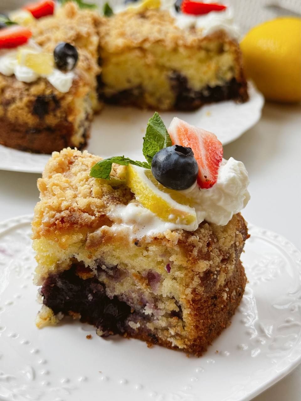 Slice of lemon yogurt cake with blueberries and whipped cream, garnished with mint, lemon and strawberry