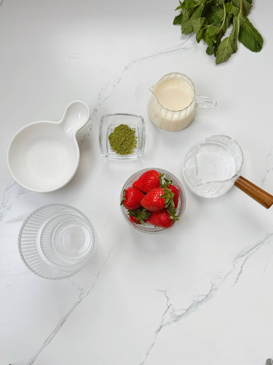 Ingredients for strawberry iced matcha latte including matcha powder, fresh strawberries, milk, and sugar