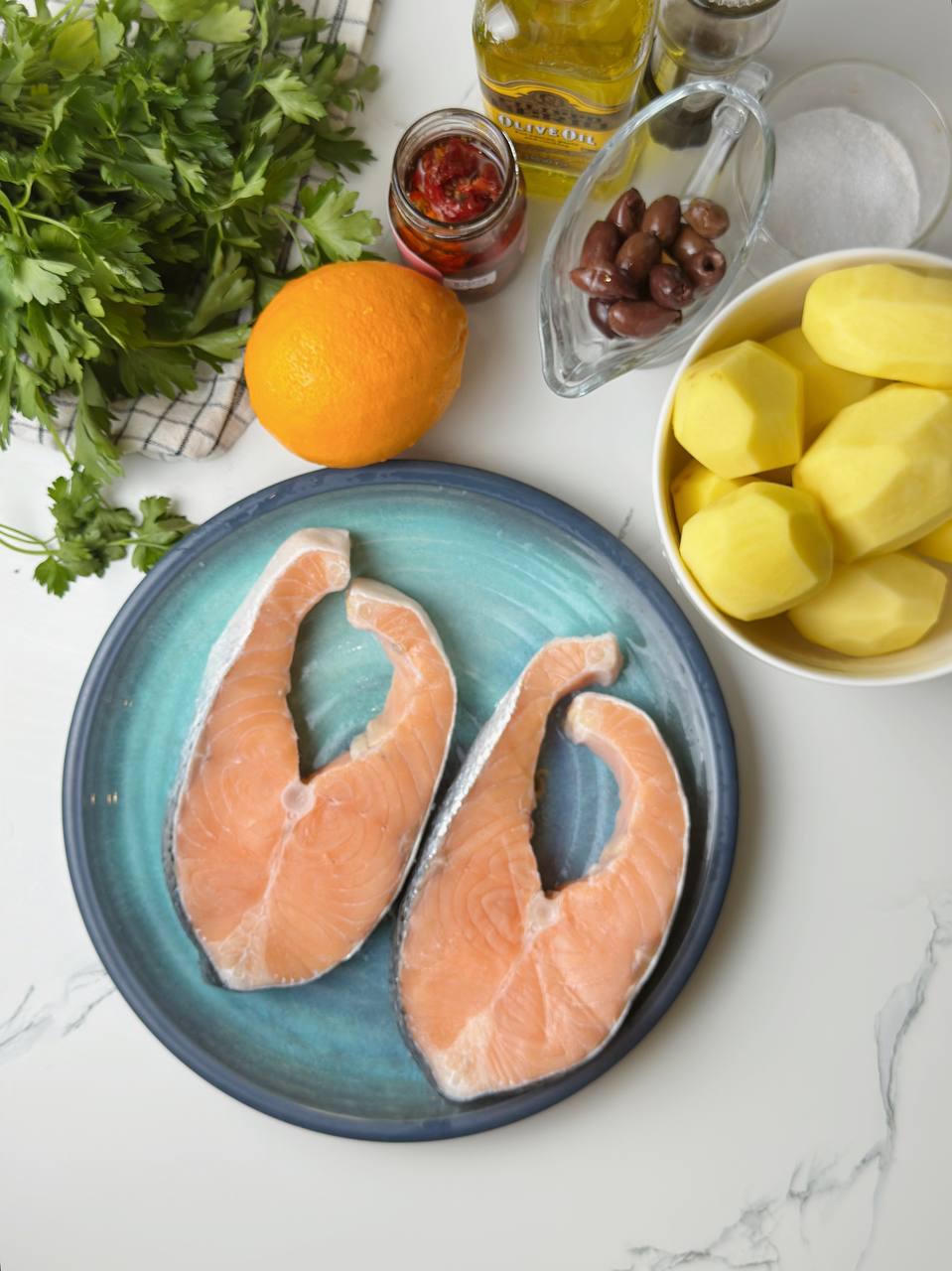 Ingredients including salmon, baby potatoes, olives, sun-dried tomatoes, parsley, and lemon on a table