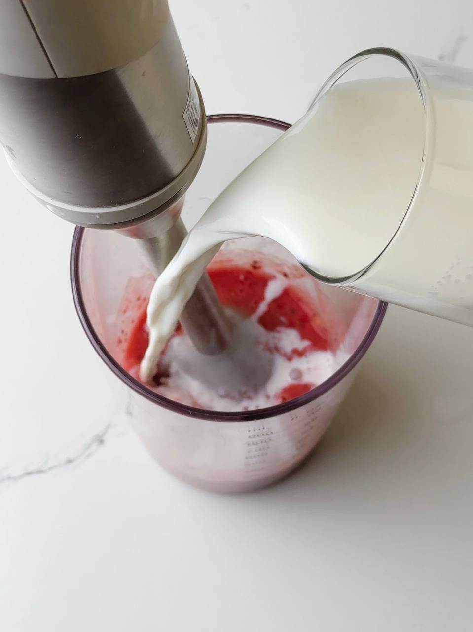 Pouring milk over fruit in blender to make creamy strawberry banana milkshake.