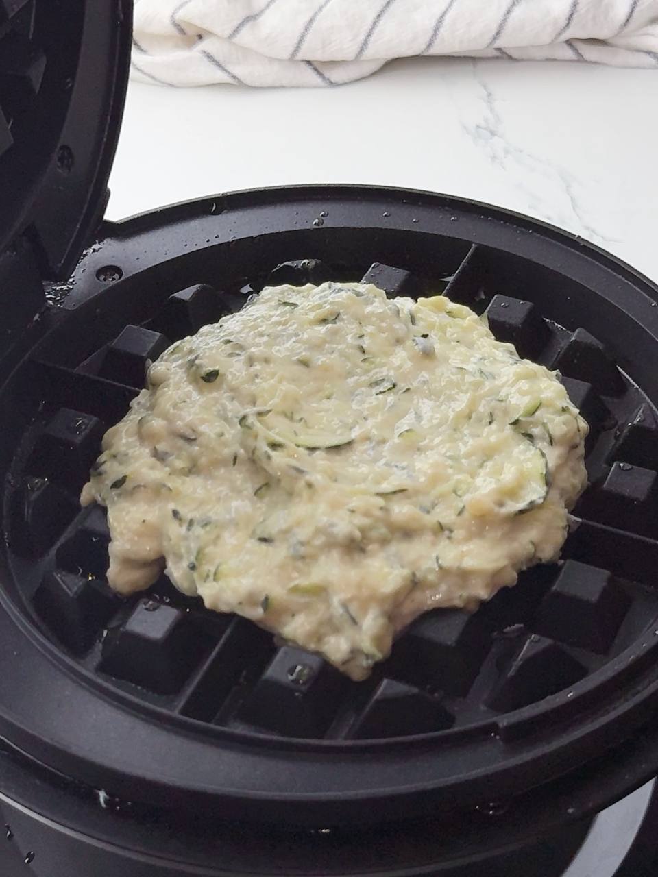 Raw zucchini waffle batter spread on hot waffle iron before cooking.
