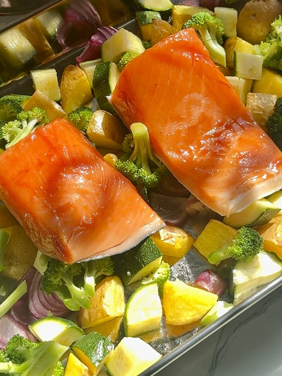 Glazed salmon fillets with potatoes, broccoli, zucchini and red onion on a baking tray