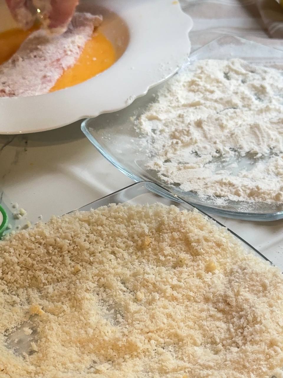 Chicken breast being dipped into beaten egg after flour coating, with panko breadcrumbs ready for the final breading step.